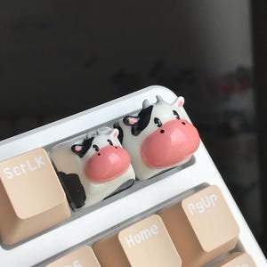 Könnte beinhalten: Zwei schwarze und weiße Kuh-Tastenkappen mit rosa Nasen, eine größer als die andere, befinden sich auf einer weißen Tastatur mit beigefarbenen Tasten, die mit "ScrLk", "Home" und "PgUp" beschriftet sind.