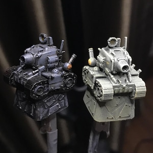 May include: Two small, gray, toy tanks with black treads and a cannon on top. The tanks are on a gray, textured base.