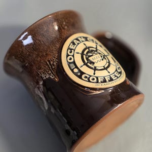 Może przedstawiać: Brązowy ceramiczny kubek z błyszczącym wykończeniem. Kubek posiada okrągły emblemat z napisem "OCEAN CITY COFFEE" i motywem marynistycznym. Uchwyt jest widoczny po prawej stronie. Kubek ma naturalną, glinianą podstawę.