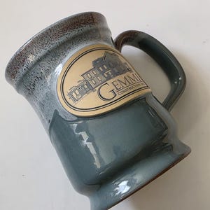 May include: A ceramic mug with a blue-grey glaze and a brown rim. The mug features a handle and an oval label with the text "GEMMI CONSTRUCTION INC" and an illustration of a building.