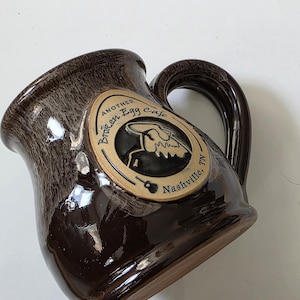 May include: A brown ceramic mug with a curved handle. The mug features a logo with the text "Another Broken Egg Cafe, Nashville, TN". The mug has a glossy finish and a textured appearance.