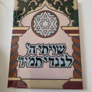May include: A decorative ceramic tile with Hebrew text and a Star of David design. The tile features a pink background with a central Star of David in light blue, surrounded by ornate green and brown borders.