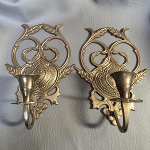 May include: Two ornate brass wall sconces. Each sconce features a candle holder, a decorative backplate with a floral design, and a curved arm. The sconces are symmetrical and have a vintage aesthetic, suitable for home decor.
