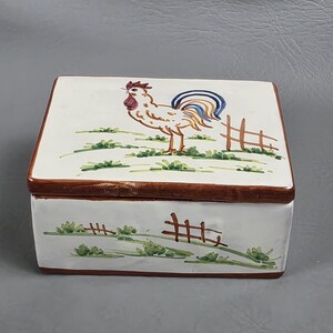 May include: A rectangular ceramic box with a lid. The box is white with a brown rim and a painted rooster design. The rooster is tan, red, and blue, standing near a small fence and green grass.
