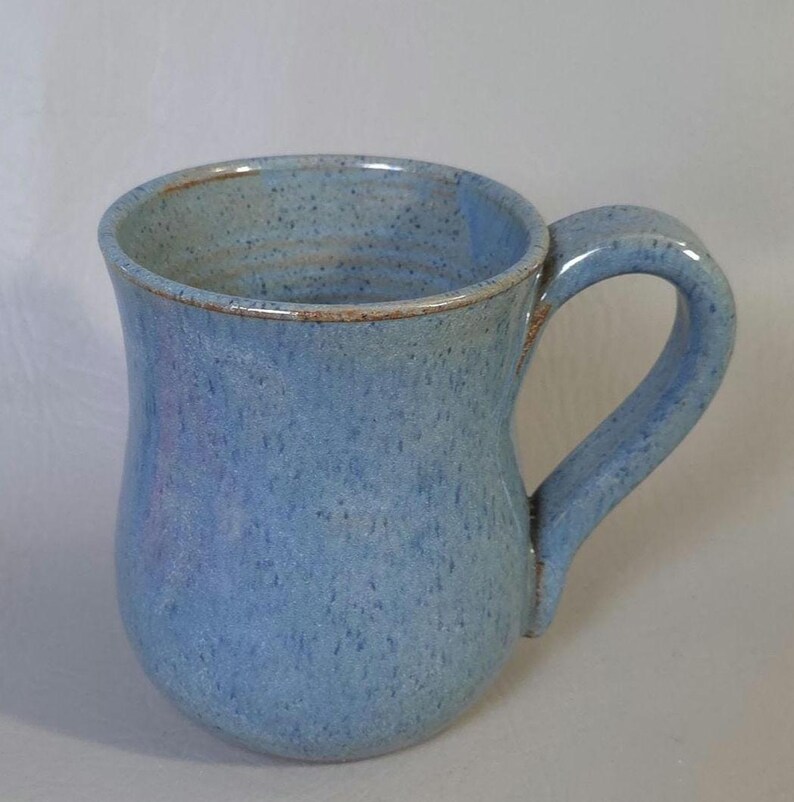 May include: A handmade ceramic mug with a light blue glaze and a curved handle. The mug has a speckled texture and a brown rim. The mug is approximately 4 inches tall and 3 inches in diameter.