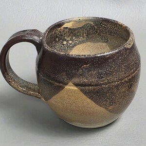 May include: A handmade ceramic mug with a rounded body and a large handle. The mug features a unique color scheme, with a dark brown exterior and a tan interior. The design includes a diagonal pattern of tan and brown. The mug is suitable for hot or cold beverages.
