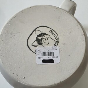 May include: A white ceramic mug with a handle, viewed from the bottom. The base features a black and white illustration of a cartoon animal and the word "OLOGIES" in a circle. A sticker with a barcode and numbers is affixed to the base.