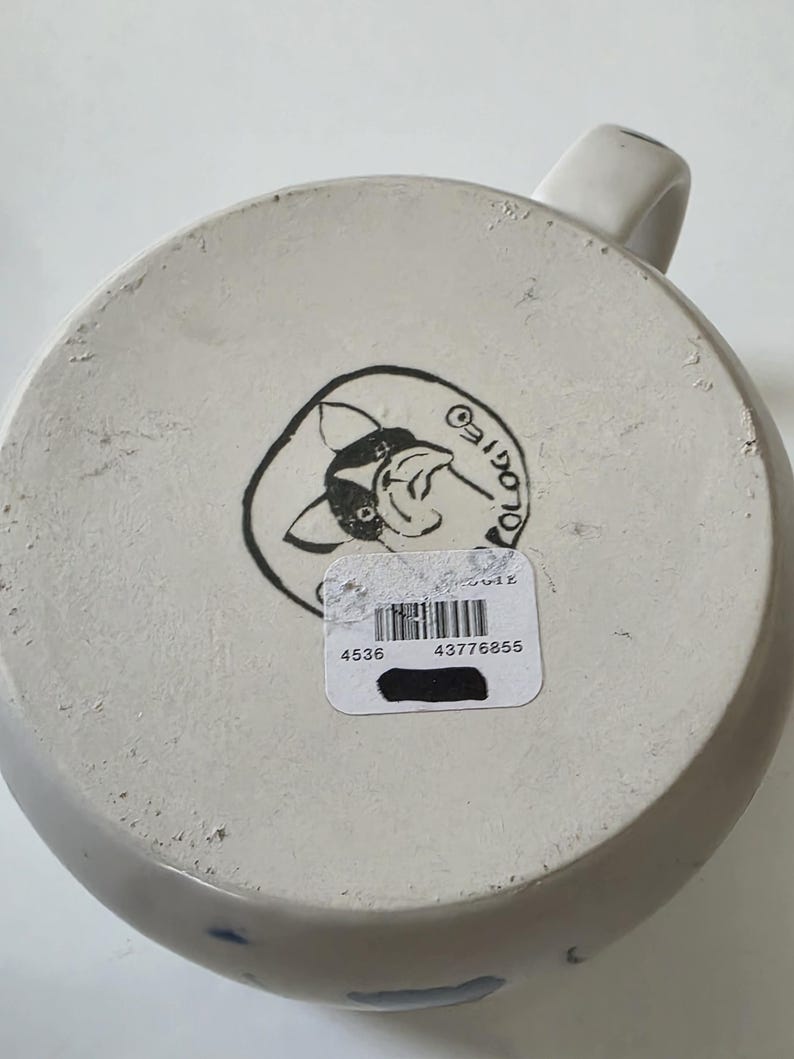May include: A white ceramic mug with a handle, viewed from the bottom. The base features a black and white illustration of a cartoon animal and the word "OLOGIES" in a circle. A sticker with a barcode and numbers is affixed to the base.