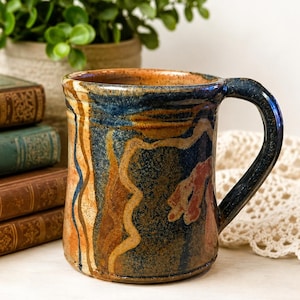 May include: A ceramic mug featuring a mottled glaze in shades of deep blue, earthy brown, and tan. The exterior surface displays abstract, wavy vertical lines and organic shapes. It has a sturdy, curved handle and a smooth, rounded rim.
