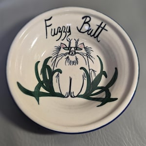 May include: A small, off-white ceramic bowl with a blue rim. The bowl features a hand-drawn illustration of a cat's rear end, with the words "Fuzzy Butt" written above. Green leaf-like designs surround the cat illustration.