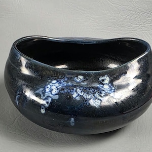 Op de afbeelding: Een donkerblauwe keramische kom met een unieke, organische vorm. De kom heeft een glanzende afwerking met witte en blauwe kristalpatronen. De binnenkant is diepzwart en contrasterend. Een decoratief en functioneel stuk voor in huis.