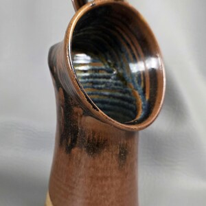May include: A handcrafted ceramic jug with a distinctive design. The jug has a brown exterior with dark brown and blue accents inside the opening. The handle is curved, and the base has a light tan colour.