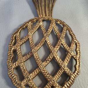 May include: A gold-colored pineapple-shaped trivet with a lattice design. The trivet has a textured surface and a detailed crown. The edges are scalloped, adding a decorative touch. This item is likely used for placing hot dishes.