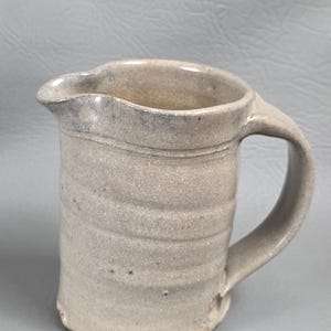 Może przedstawiać: Ręcznie wykonany, beżowy ceramiczny dzbanek z uchwytem i dzióbkiem. Dzbanek ma teksturowaną powierzchnię i nieco ciemniejszą krawędź. Projekt jest prosty i rustykalny, odpowiedni do serwowania napojów lub jako element dekoracyjny.