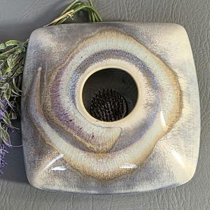 May include: Square ceramic vase with a central circular opening. The vase has a swirling pattern of cream, purple, and brown hues. The opening contains a black floral frog. Decorative lavender sprigs are placed near the vase.