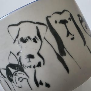 May include: White ceramic mug with a blue rim, featuring black line drawings of various dog breeds. The mug's design includes several dog illustrations, showcasing different shapes and sizes.