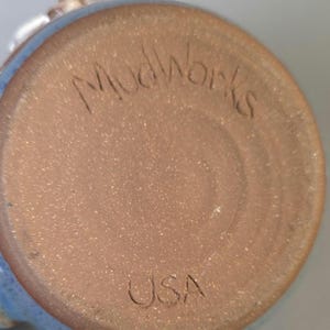 May include: Close-up of the bottom of a ceramic mug. The base is a speckled brown with the etched text "Mudworks" and "USA". The rim of the mug is a light blue color.