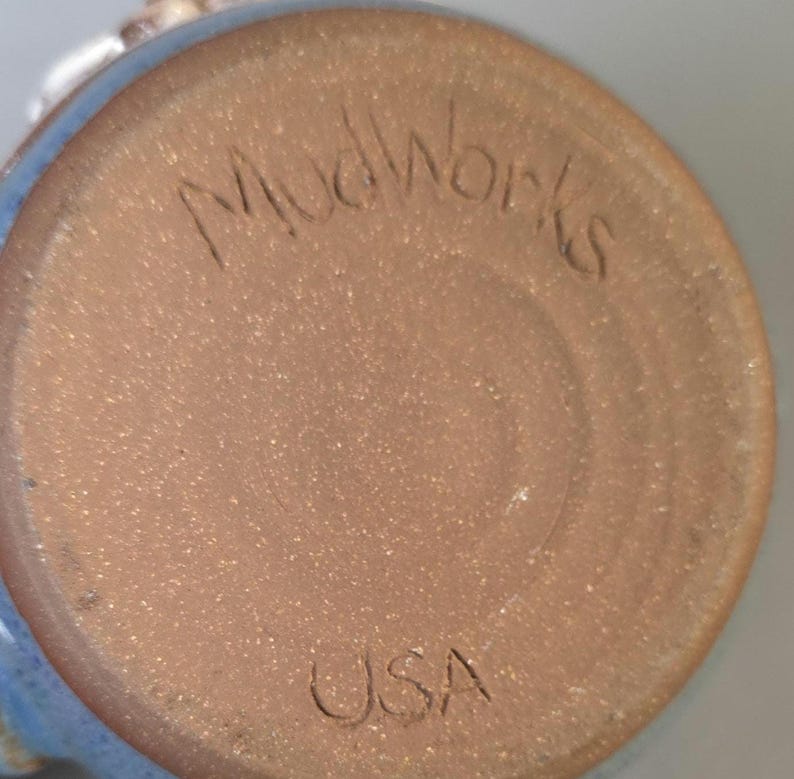 May include: Close-up of the bottom of a ceramic mug. The base is a speckled brown with the etched text "Mudworks" and "USA". The rim of the mug is a light blue color.