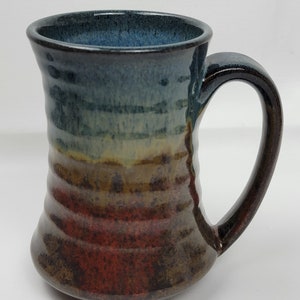 May include: A handmade ceramic mug with a blue, green, yellow, and brown glaze. The mug has a wide base and a flared top.