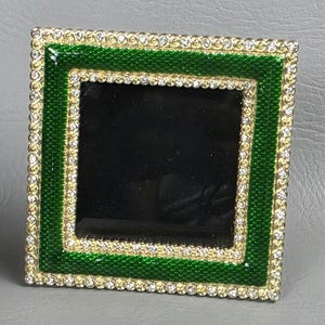 May include: Square picture frame with a green enamel border and a gold inner border. The frame is encrusted with small, clear rhinestones around the edges. The centre is black.