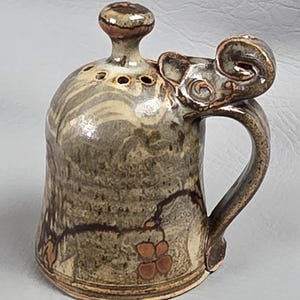 May include: A handcrafted ceramic mug with a unique elephant head handle. The mug features a speckled brown and tan glaze with a decorative floral design. The lid has a small knob and several holes. A charming and functional piece.