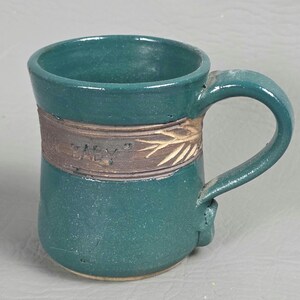 May include: A handmade teal ceramic mug with a rounded body and a large handle. The mug features a brown band with the word "BABY" and a leaf design. The mug is approximately 10 cm tall.