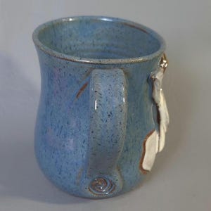 May include: A handmade ceramic mug with a light blue speckled glaze. The mug features a curved handle and a white and brown decorative element on the side. The mug is a unique piece of pottery.