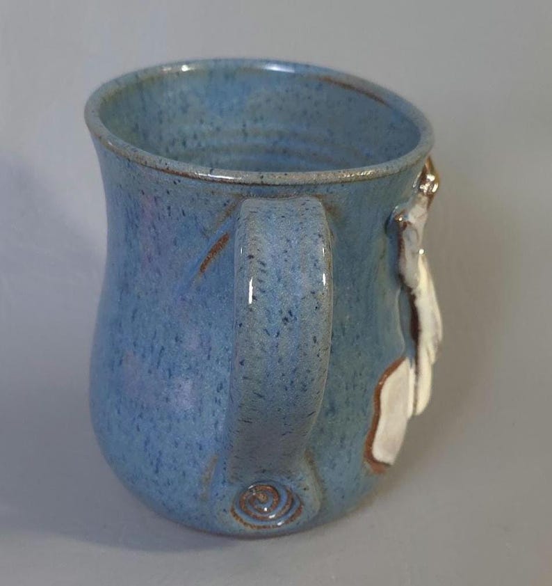May include: A handmade ceramic mug with a light blue speckled glaze. The mug features a curved handle and a white and brown decorative element on the side. The mug is a unique piece of pottery.
