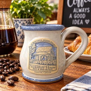 Puede incluir: Una taza de cerámica con borde y base de color azul moteado, que presenta una calcomanía central de una casa con el texto 'IN BEAUTIFUL INDIAN VALLEY', 'Yorkshire House BED & BREAKFAST' y 'Greenville, California' sobre un fondo crema.