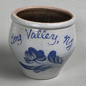 Puede incluir: Una olla de cerámica artesanal de color blanquecino con un diseño floral azul y la inscripción "Long Valley, NJ". El borde es de color rosa claro y el interior es marrón oscuro. Esta pieza rústica es ideal para la decoración del hogar.