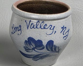 Puede incluir: Una olla de cerámica artesanal de color blanquecino con un diseño floral azul y la inscripción "Long Valley, NJ". El borde es de color rosa claro y el interior es marrón oscuro. Esta pieza rústica es ideal para la decoración del hogar.