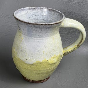 May include: A handmade ceramic mug with a large handle. The mug features a color gradient, transitioning from a light gray rim to white, and then to a yellow base. The mug has a rustic, textured appearance.