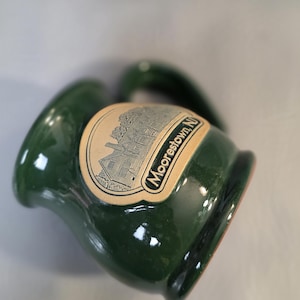 May include: A dark green ceramic mug with a handle. The mug has a glossy finish and a label that reads "Moorestown, NJ" with an illustration of a building. The mug is tilted, showing its shape and design.