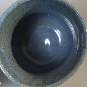 May include: A handmade ceramic bowl with a speckled blue glaze. The bowl has a rounded shape with a wide opening and a smooth interior. The exterior has a textured, speckled appearance.