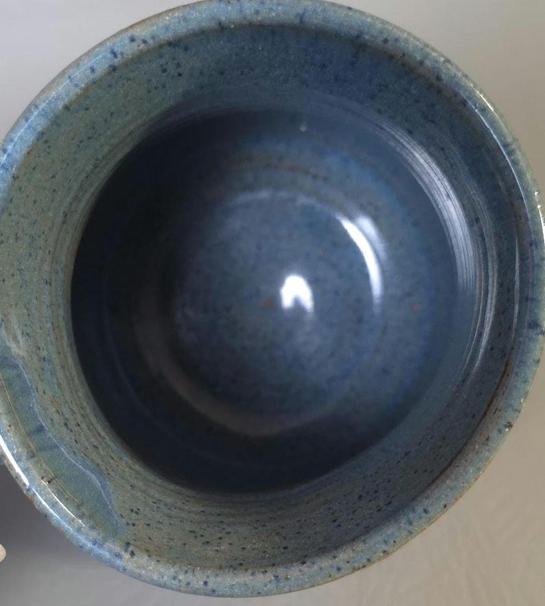 May include: A handmade ceramic bowl with a speckled blue glaze. The bowl has a rounded shape with a wide opening and a smooth interior. The exterior has a textured, speckled appearance.