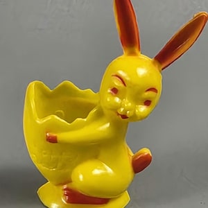 Może przedstawiać: Żółta ceramiczna figurka zajączka wielkanocnego trzymająca żółtą skorupkę jajka. Zajączek ma czerwone akcenty na uszach, nosie i stopach. Figurka jest przedmiotem dekoracyjnym, prawdopodobnie na Wielkanoc.