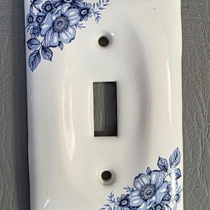 Pode incluir: Tampa de interruptor de luz de cerâmica branca com design floral azul. A tampa tem uma abertura retangular para o interruptor e dois orifícios para parafusos. Padrões florais azuis adornam os cantos superior esquerdo e inferior direito.