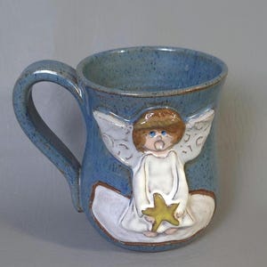 May include: A blue ceramic mug with a handle, featuring a raised relief of an angel holding a yellow star. The angel has brown hair, white wings, and a white robe. The mug has a speckled finish.