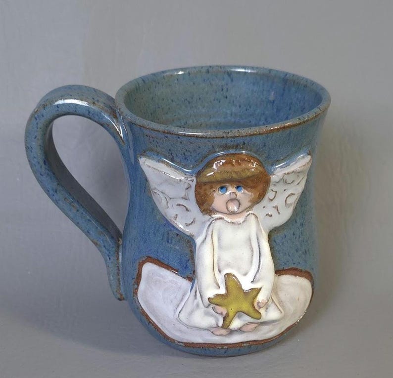 May include: A blue ceramic mug with a handle, featuring a raised relief of an angel holding a yellow star. The angel has brown hair, white wings, and a white robe. The mug has a speckled finish.