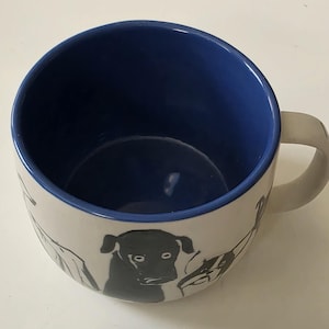 May include: White ceramic mug with a blue interior and a white handle. The exterior features black line drawings of dogs. The mug is designed for hot beverages and has a simple, modern aesthetic.