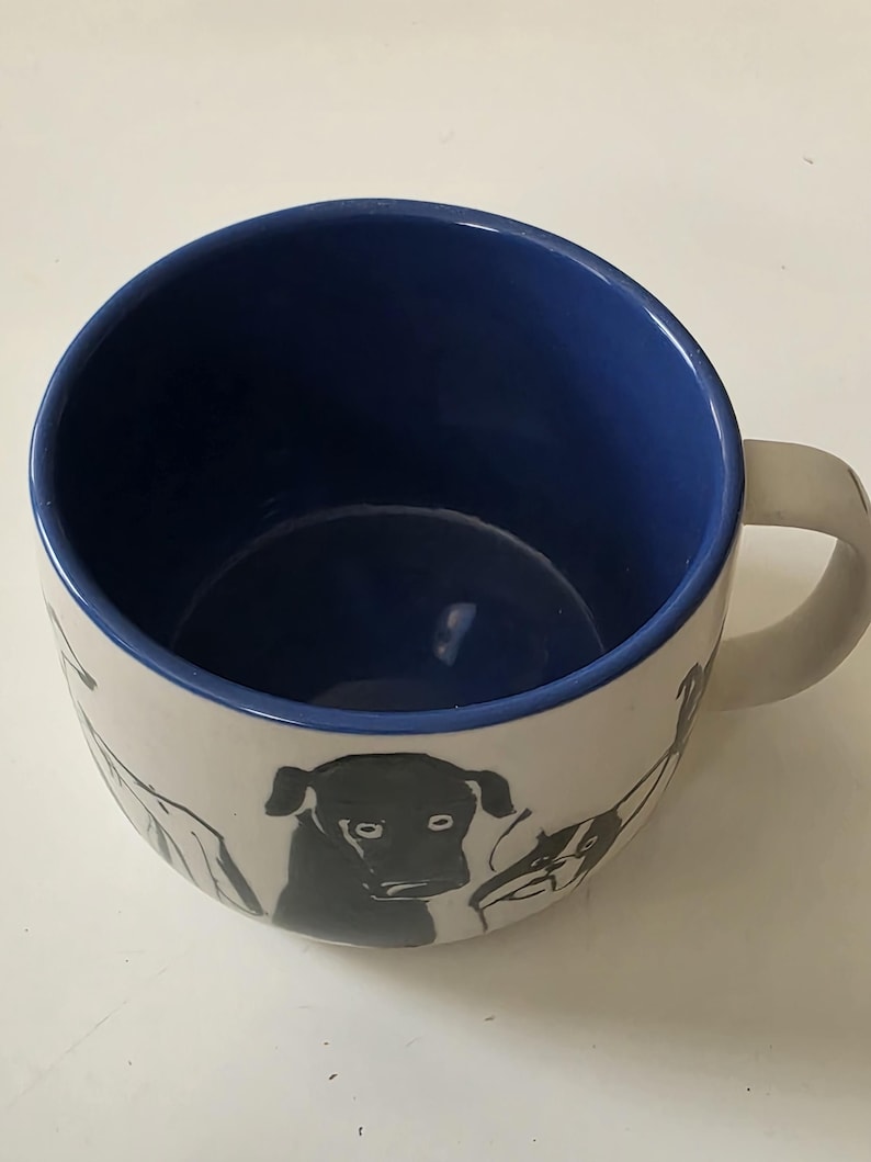 May include: White ceramic mug with a blue interior and a white handle. The exterior features black line drawings of dogs. The mug is designed for hot beverages and has a simple, modern aesthetic.