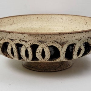 May include: A speckled ceramic bowl with a pedestal base. The bowl has a light beige interior and a dark brown rim. The sides feature a repeating pattern of interlocking circles, creating a decorative, open design. Suitable for serving or display.