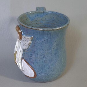 May include: A handmade ceramic mug with a light blue speckled glaze. The mug features a sculpted white and brown floral design on the side and a small handle. The mug is approximately 4 inches tall.
