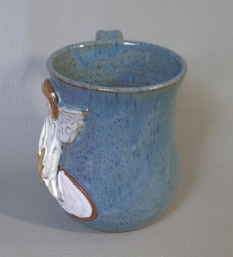 May include: A handmade ceramic mug with a light blue speckled glaze. The mug features a sculpted white and brown floral design on the side and a small handle. The mug is approximately 4 inches tall.