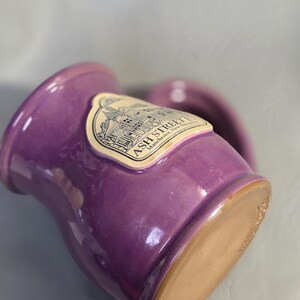 May include: A purple ceramic mug with a handle. The mug has a glossy finish and a light brown base. A label on the side reads "ASH STREET" with an illustration of a building. The mug is likely handmade.