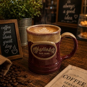 May include: A ceramic mug with a two-tone burgundy and tan glaze, featuring a central oval emblem that reads C.F. Martin & Co. EST. 1833. The mug is filled with coffee topped with latte art, positioned near a small chalkboard sign and coffee beans.