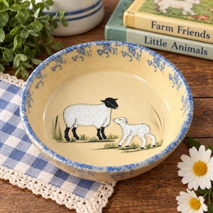 May include: A round ceramic bowl with a cream-coloured base and a speckled finish. The interior features a hand-painted illustration of a black-faced sheep and a lamb standing in green grass. The rim has a blue sponge-painted floral pattern.