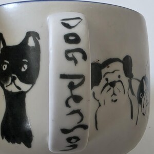 May include: White ceramic mug with black dog illustrations and the text "Dog Person" on the handle. The mug features various dog breeds in a simple, illustrative style, with a blue rim.