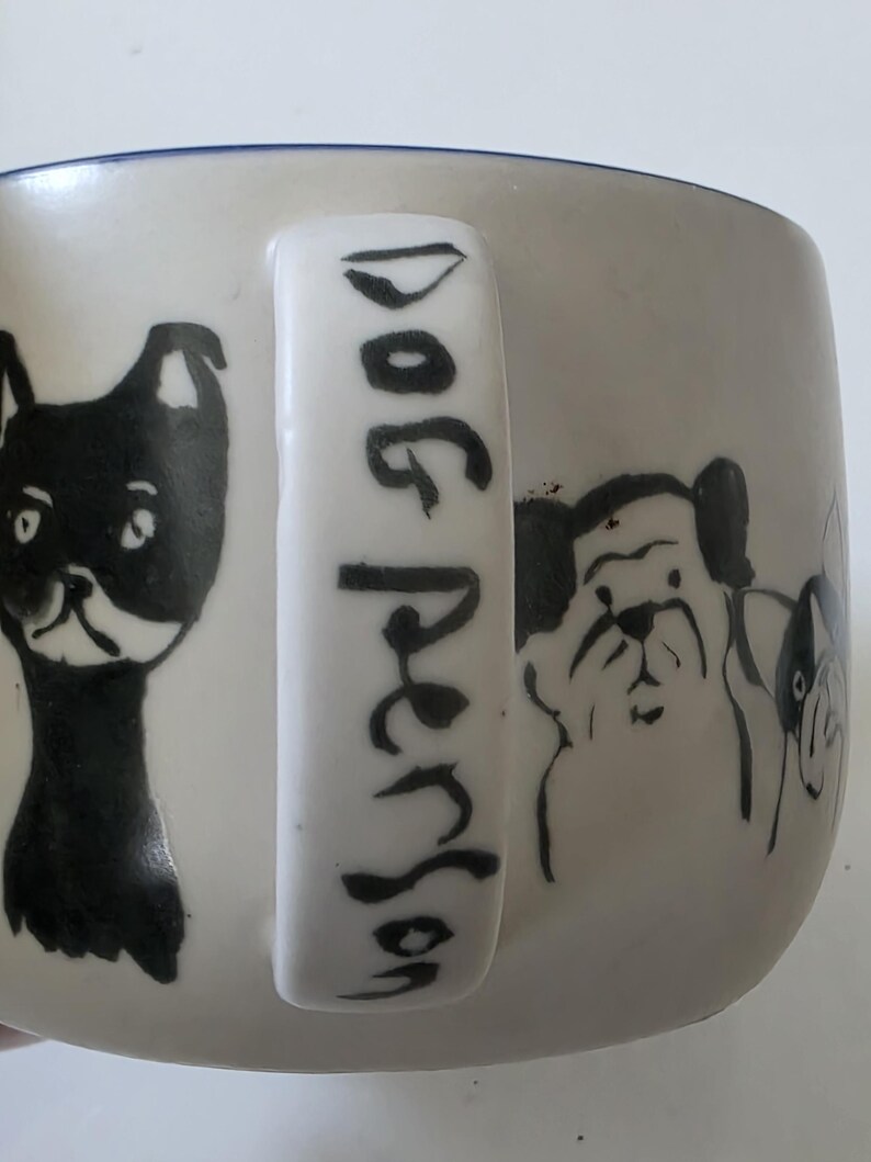 May include: White ceramic mug with black dog illustrations and the text "Dog Person" on the handle. The mug features various dog breeds in a simple, illustrative style, with a blue rim.