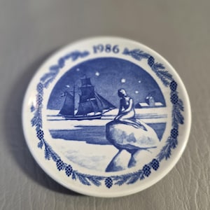May include: A round, white plate with a blue illustration of a mermaid on a rock, a sailing ship, and the year 1986. The plate has a blue border with pine cone and leaf designs. A decorative piece.